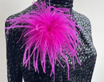 Puede incluir: Un broche de plumas rosa intenso sujeto a una blusa negra de cuello alto con un sutil estampado floral gris. El texto hot pink aparece sobre el accesorio circular de plumas, que presenta plumas largas, finas y radiantes.