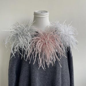 May include: A dark gray sweater featuring a decorative collar made of layered feathers in shades of white, light gray, and soft pink. The feathers are interspersed with thin, metallic silver strands that add a reflective quality to the texture.