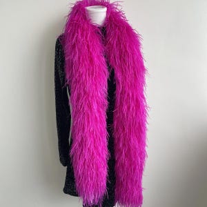 Puede incluir: Una boa de plumas de color rosa intenso drapeada sobre un vestido negro de manga larga con un sutil patr&oacute;n texturizado. La boa est&aacute; compuesta por plumas largas, vaporosas y voluminosas que crean una apariencia densa alrededor del cuello.