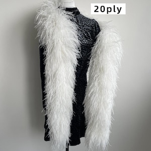 Puede incluir: Una boa de plumas blanca larga con un grosor de 20ply, drapeada sobre una prenda negra texturizada de cuello alto. El texto 20ply es claramente visible en la esquina superior derecha de la imagen, indicando la densidad de las plumas blancas.