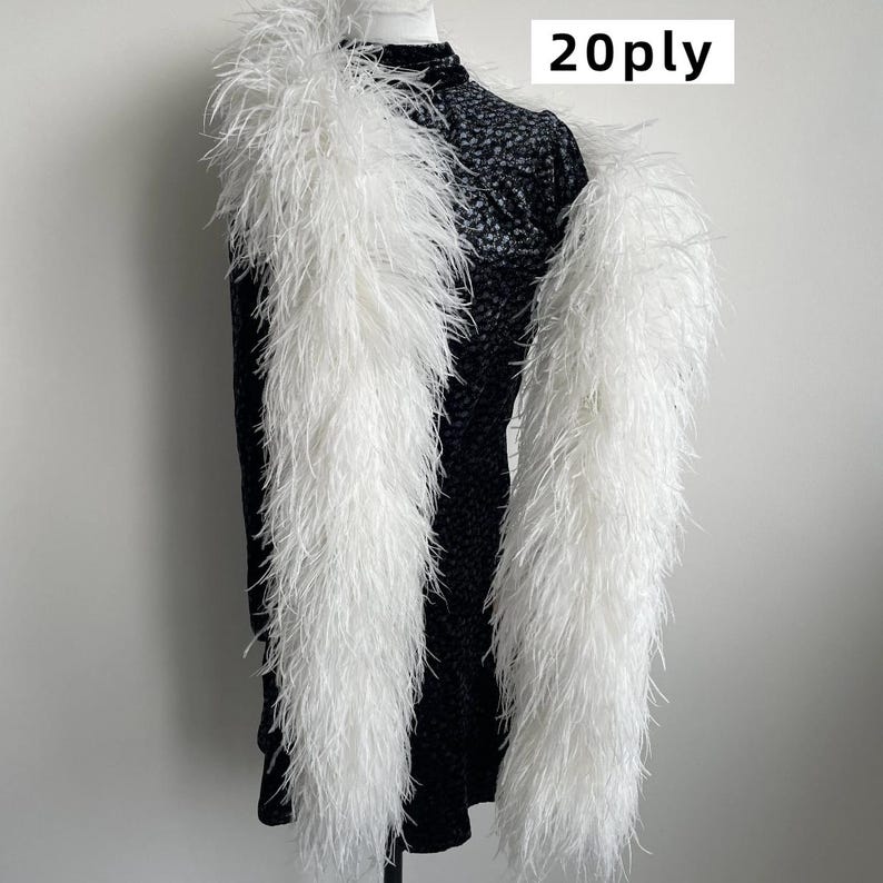 Puede incluir: Una boa de plumas blanca larga con un grosor de 20ply, drapeada sobre una prenda negra texturizada de cuello alto. El texto 20ply es claramente visible en la esquina superior derecha de la imagen, indicando la densidad de las plumas blancas.