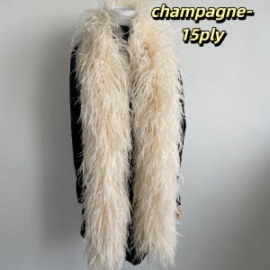 Puede incluir: Un largo y esponjoso boa de plumas color champ&aacute;n drapeado sobre una prenda negra. El boa presenta una textura densa de m&uacute;ltiples capas y lleva el texto champagne-15ply en la esquina superior derecha de la imagen.