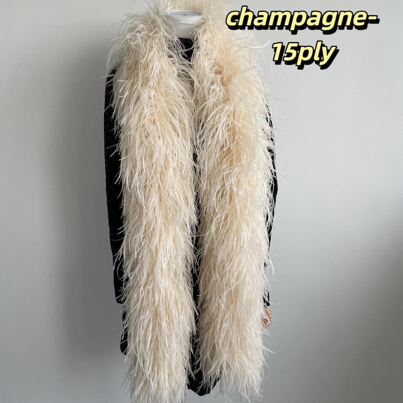 Puede incluir: Un largo y esponjoso boa de plumas color champ&aacute;n drapeado sobre una prenda negra. El boa presenta una textura densa de m&uacute;ltiples capas y lleva el texto champagne-15ply en la esquina superior derecha de la imagen.