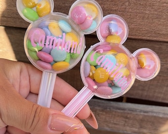 May include: Two clear plastic Mickey Mouse-shaped containers with light pink handles, filled with colourful candy-coated chocolates. Each container features the name Camiland printed in pink script across the centre of the circular clear dome.