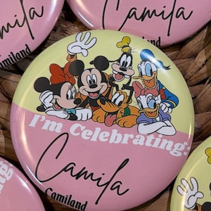 May include: A round pinback button featuring a split yellow and pink background. The design includes Mickey Mouse, Minnie Mouse, Goofy, Pluto, Donald Duck, and Daisy Duck. The text reads I'm Celebrating: Camila Camiland in black script.