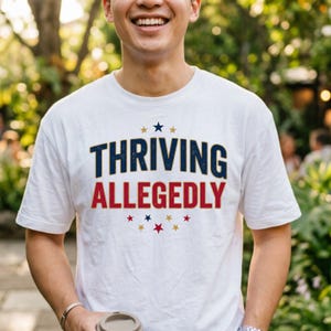 May include: A white short-sleeved t-shirt featuring the text THRIVING in dark blue and ALLEGEDLY in red. The design includes small navy blue and gold stars arranged in an arc above and below the text, with a distressed texture on the letters.
