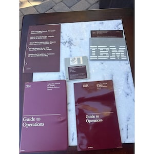Może przedstawiać: Zestaw materiałów do karty sieciowej IBM Token-Ring PC Adapter, zawierający dwie bordowe instrukcje Guide to Operations, dyskietkę 3,5 cala w szarej kopercie z logo IBM, luźną dyskietkę oraz bordowy arkusz informacyjny z tekstem w wielu językach.