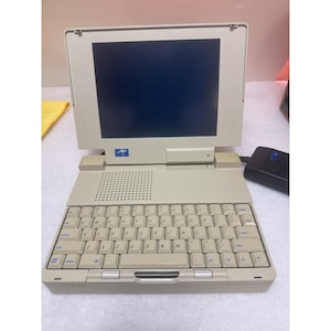 Può includere: Un computer portatile beige vintage con schermo scuro, tastiera QWERTY e un piccolo logo a forma di canguro blu sulla cornice del display. Il dispositivo include una griglia per altoparlanti sopra i tasti e un alimentatore nero sulla destra.