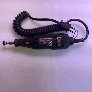 May include: A black Dremel Moto-Tool Model 395 rotary tool with a coiled black power cord and plug. The tool features a variable speed switch, an orange power button, and a metal hanging loop at the rear. The label displays Dremel Moto-Tool Model 395.