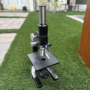 Puede incluir: Un microscopio negro Bausch & Lomb con un ocular plateado, dos lentes objetivo y una platina plana con dos clips de metal. El instrumento incluye perillas de ajuste plateadas en el lateral y un espejo circular situado bajo la platina.