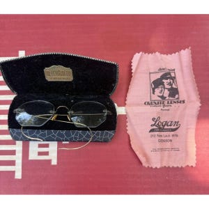 Peut inclure: Lunettes sans monture avec des branches en métal doré fin dans un étui noir avec une plaque dorée indiquant Dr. J. Logan O.D. Denison, Iowa. À côté de l'étui se trouve un chiffon rose avec le texte Cruxite Lenses, Logan for Glasses, 212 New Laub Bldg.