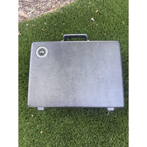 Puede incluir: Un maletín rígido rectangular de color gris con superficie texturizada, asa superior negra y dos pequeños pies en la base. Un emblema circular con un gráfico de tractor y el texto 1899 se encuentra en la esquina superior izquierda del maletín.