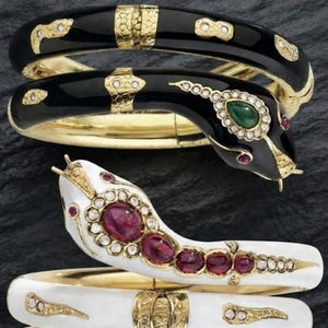 Puede incluir: Dos pulseras articuladas con forma de serpiente hechas de oro. Una pulsera presenta esmalte negro con una cabeza de piedra verde y detalles de diamantes. La otra pulsera presenta esmalte blanco con una serie de piedras rojas y diamantes.