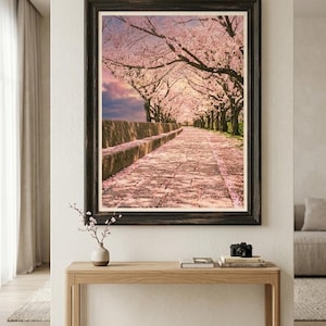 以下が含まれることがあります： 夕焼け空の下、満開の桜並木と石畳の小道が描かれた額装アートプリント。地面に散らばるピンクの花びら、暗い木の枝、石垣が特徴で、ダークウッドのフレームに収められています。