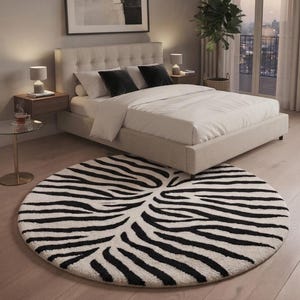Op de afbeelding: Een rond vloerkleed met een contrastrijk zwart-wit zebrastrepenpatroon. Het kleed heeft een dikke, getextureerde pool en ligt op een lichte houten vloer voor een bed met een beige gecapitonneerd hoofdeinde.