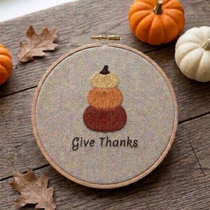 May include: A circular wooden embroidery hoop containing a gray fabric panel with a hand-stitched design of three stacked pumpkins in shades of yellow, orange, and brown. Below the pumpkins, the text Give Thanks is embroidered in black thread.