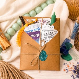 May include: A brown paper envelope tied with twine and a teal wax seal, containing cards with sun illustrations and text reading Charge Crystals, Burn Sage, and Yogi. The envelope is surrounded by light green beads, a candle, and crystals.
