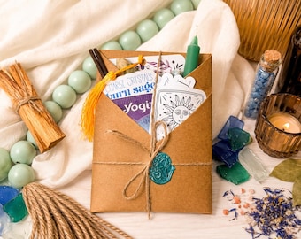 May include: A brown paper envelope tied with twine and a teal wax seal, containing cards with sun illustrations and text reading Charge Crystals, Burn Sage, and Yogi. The envelope is surrounded by light green beads, a candle, and crystals.