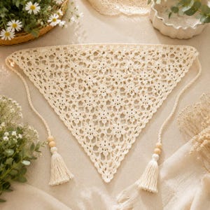 May include: A triangular off-white crochet bandana featuring an intricate floral lace pattern. The accessory includes two long braided ties, each finished with a single wooden bead and a matching off-white yarn tassel at the ends.