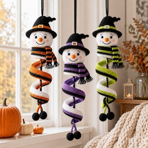 May include: Three hanging crochet snowman ornaments wearing witch hats. Each snowman has a striped scarf and a long, spiralling body in orange and white, purple and black, or lime green and white. They hang from black cords with small pom-poms.