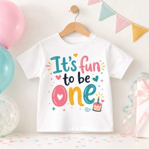 May include: A white short-sleeved t-shirt featuring the colorful text It's fun to be one in shades of teal, pink, dark blue, and yellow. The design includes small heart icons, star shapes, and a small birthday cake illustration with a single candle.
