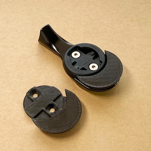 May include: A black bicycle computer mount featuring a glossy arm and a matte circular head with two silver screws. The mount includes a separate, detached black circular base piece with two screw holes, both components showing a textured finish.
