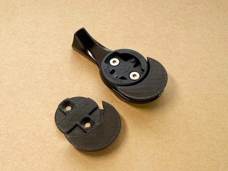 May include: A black bicycle computer mount featuring a glossy arm and a matte circular head with two silver screws. The mount includes a separate, detached black circular base piece with two screw holes, both components showing a textured finish.