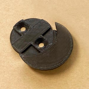 May include: A black 3D printed circular component featuring a crescent-shaped extension on one side and a central cross-shaped support structure with two circular mounting holes positioned within the recessed areas of the main circular body.