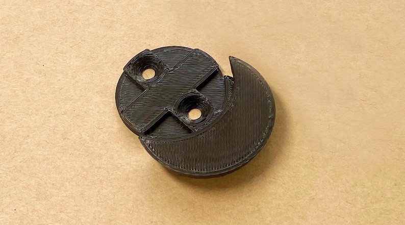 May include: A black 3D printed circular component featuring a crescent-shaped extension on one side and a central cross-shaped support structure with two circular mounting holes positioned within the recessed areas of the main circular body.