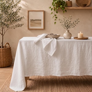 Peut inclure: Une nappe en lin blanc drapée sur une table rectangulaire. Le tissu présente un tissage visible et retombe sur les côtés. Une serviette blanche pliée repose sur la surface de la table, à côté d'un bol en céramique, d'un vase avec des feuilles.