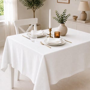 May include: A square white tablecloth draped over a dining table, set with two white plates, folded white napkins, gold-toned forks and knives, a clear ribbed glass, a tall white taper candle in a brass holder, and a small amber glass votive.