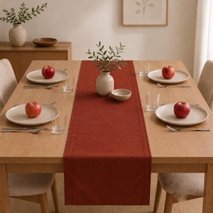 Puede incluir: Un camino de mesa rectangular de color óxido centrado en una mesa de comedor de madera clara. La mesa está puesta con cuatro platos blancos, cada uno con una manzana roja, cuatro vasos transparentes y cubiertos de plata. Un jarrón de cerámica con hojas verdes se encuentra en el centro.