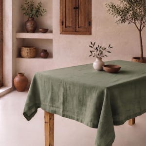 May include: A rectangular sage green tablecloth with a visible fabric texture draped over a wooden table. On the table sits a small white ceramic vase containing a green leafy branch and a plain brown wooden bowl. The cloth has a hemmed edge.