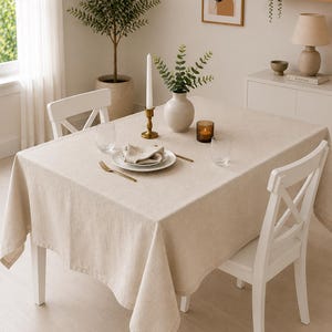 Peut inclure: Une nappe rectangulaire en lin beige recouvrant une table à manger. La table est dressée avec une assiette blanche, une serviette pliée, une fourchette dorée, deux verres transparents, un bougeoir en laiton et un vase avec du feuillage vert.