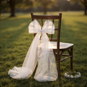 May include: A wooden chair adorned with a large white tulle bow and flowing fabric. A small floral wreath rests on the grass below. The scene is set outdoors with soft sunlight.