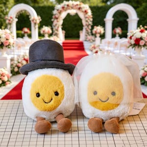 May include: Two plush egg-shaped toys with soft white bodies and yellow centers. One toy wears a dark gray felt top hat, while the other wears a sheer white veil. Both have small black eyes, embroidered smiles, and dangling brown corduroy legs.