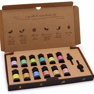 Könnte beinhalten: Eine braune Pappschachtel mit zwölf kleinen ätherischen Ölflaschen aus Glas mit bunten Etiketten und zwei schwarzen Pipetten. Der Innendeckel zeigt den Text a guide to essential oils mit Symbolen und Bezeichnungen für die Düfte citrus, minty, floral, herbaceous, spicy und woody.