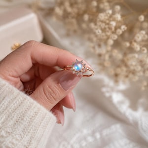 May include: A rose gold ring featuring a central round iridescent moonstone cabochon surrounded by a halo of small clear sparkling stones arranged in a sunburst pattern. The thin metal band is polished and smooth, showcasing a delicate design.