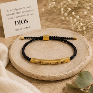 May include: A black braided cord bracelet featuring a curved gold-tone metal bar engraved with the text DIOS TIENE EL CONTROL. The bracelet includes a sliding gold-tone metal adjustment bead and matching end caps on the cord tips.