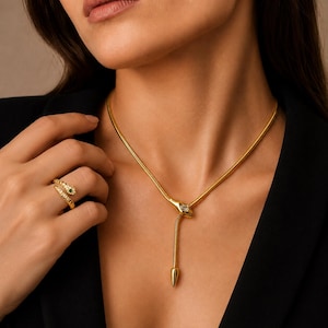 May include: A gold snake-themed necklace featuring a flexible chain with a central snake head motif and a dangling tail end, paired with a matching gold snake ring adorned with small white stones and a single green stone on the head.