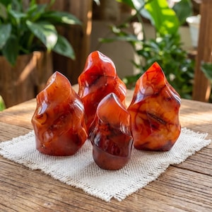 May include: Four polished carnelian crystal carvings shaped like flames. Each piece features a smooth, glossy surface with deep orange, red, and brownish-red colour variations, showcasing natural banding and translucent qualities in a flame-like form.