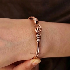 May include: A copper-coloured wire bangle bracelet featuring a hook-and-eye style clasp mechanism. The design consists of two parallel metal wires secured by tightly coiled wire wraps at the closure point. The text Jewelleryvilla is visible.