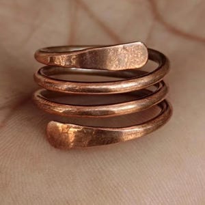 May include: A copper-coloured metal ring featuring a spiral design with three distinct coils. The surface of the metal has a hammered, textured finish, and the two ends of the spiral are flattened and rounded, creating a continuous wrap-around band.