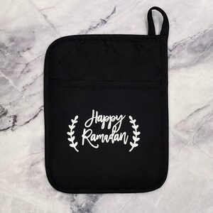 May include: Black oven mitt with a white embroidered design that says "Happy Ramadan" with a wreath of leaves around the text.
