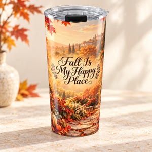 Puede incluir: Un vaso de acero inoxidable con tapa transparente que presenta un paisaje otoñal con follaje naranja y rojo, un camino de piedra y calabazas. El texto "Fall Is My Happy Place" está impreso en letra cursiva negra en el centro.