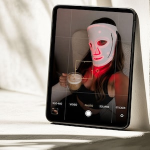 May include: A black tablet displaying a camera interface with a screen showing a person wearing a white LED light therapy mask that emits a red glow. The interface includes text labels for SLO-MO, VIDEO, PHOTO, SQUARE, and STICKER modes.