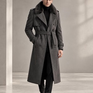 Puede incluir: Una gabardina gris carbón de doble botonadura con cuello de piel oscura, cinturón a juego con hebilla, charreteras en los hombros y puños abotonados. El abrigo se lleva sobre un suéter de cuello alto negro y pantalones negros.