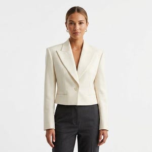 Peut inclure: Un blazer court de couleur crème doté d'une fermeture à un seul bouton, d'épaules structurées et de larges revers, associé à un pantalon gris anthracite foncé. Le blazer a des manches longues et une coupe ajustée s'arrêtant à la taille.