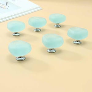 May include: A set of six cabinet knobs, each featuring a rounded, frosted light blue glass top with a smooth, pebble-like shape. Each knob is mounted on a polished silver-tone metal base, providing a clean, reflective contrast to the matte glass.