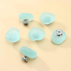 May include: A set of six cabinet knobs featuring smooth, frosted, light seafoam green glass tops with a rounded, irregular shape. Each knob is attached to a polished silver-tone metal base, providing a clean and functional hardware component.